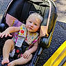 baby, child, car_seat, harness, blonde_hair, outdoor, pavement, yellow_curb, clothing, seat_belt, infant, person, cute, sitting, foot, hand, sunlight, colorful, striped, safety