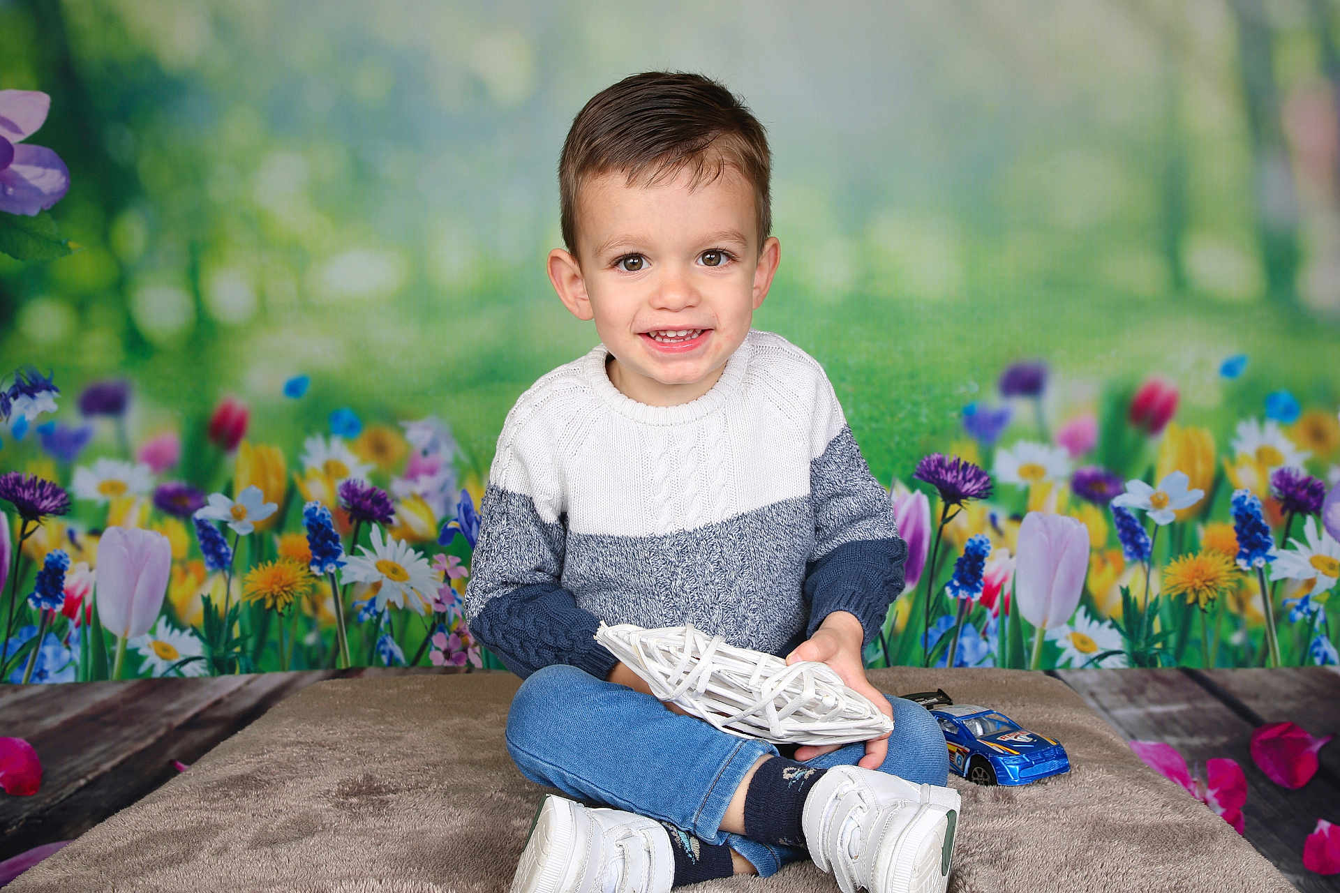 Naïm participe au concours pour gagner de l'argent avec cette photo : child, boy, smiling, sweater, jeans, white_shoes, toy_car, flower_background, indoor, portrait, cross_legged, holding_object, happy, cute, young, casual_clothing, colorful, decorative, playful, studio