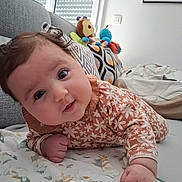 Lilia a rejoint le concours — aidez-le/la à gagner de superbes lots ! baby, infant, child, face, cute, curly_hair, floral_clothing, blanket, couch, pillow, plush_toys, indoor, window, patterned_fabric, smiling, portrait, lying_down, home, cozy, playful