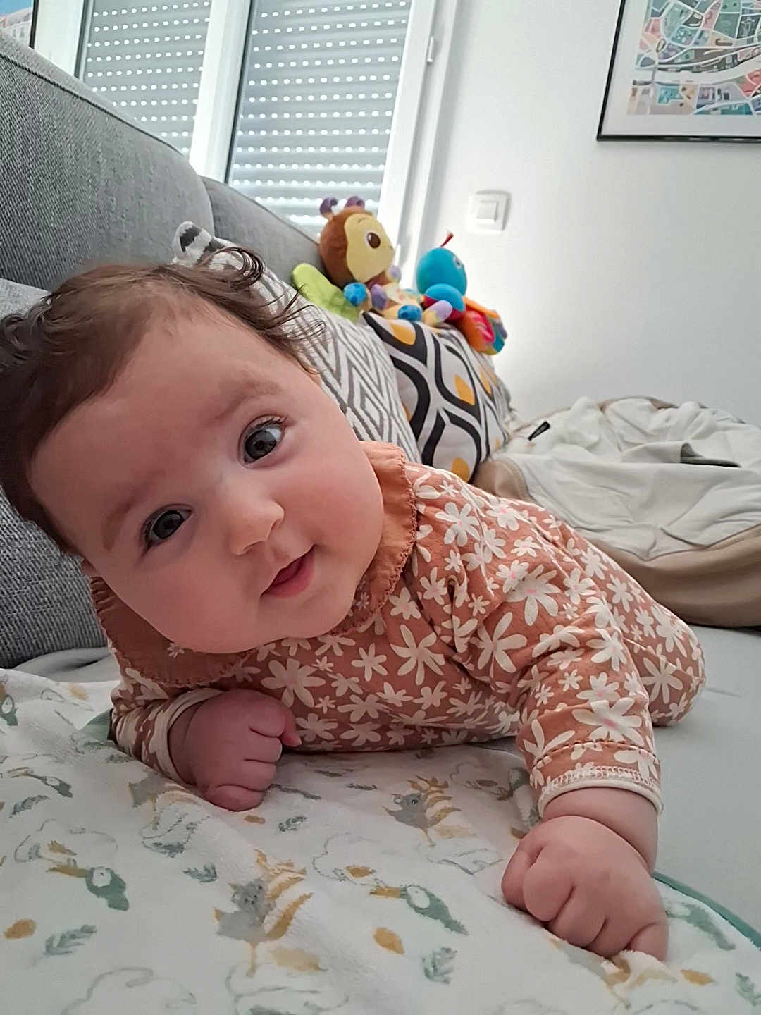 Lilia a rejoint le concours — aidez-le/la à gagner de superbes lots ! baby, infant, child, face, cute, curly_hair, floral_clothing, blanket, couch, pillow, plush_toys, indoor, window, patterned_fabric, smiling, portrait, lying_down, home, cozy, playful