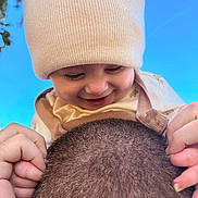 Gabriel is registered to the contest to win money with this photo: baby, child, infant, smiling, laughing, knit_hat, winter_clothing, romper, adult, head, hair, buzz_cut, hands, outdoor, blue_sky, trees, close_up, portrait, family, playful
