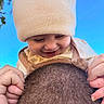 baby, child, infant, smiling, laughing, knit_hat, winter_clothing, romper, adult, head, hair, buzz_cut, hands, outdoor, blue_sky, trees, close_up, portrait, family, playful