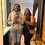 two_people, smiling, mirror_selfie, dog, pet, indoor, casual_clothing, glasses, headphones, hoodie, dress, handbag, flooring, door, cabinet, lighting, wooden_shelves, reflection, happy, young_adults