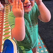 Lucas is registered to the contest to win money with this photo: toddler, child, sunglasses, green_romper, dinosaur_print, smiling, indoor, colorful_floor, star_pattern, blue_sneaker, adult, people, casual_clothing, footwear, playful, happy, short_hair, standing, person, family