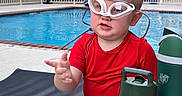 Lucas is registered to the contest to win money with this photo: blue_shorts, building, child, daylight, expression, fence, goggles, hand_gesture, lights, outdoor, pool, red_shirt, sitting, summer, swimwear, table, toddler, tree, water, water_bottle