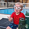 Lucas is registered to the contest to win money with this photo: toddler, child, goggles, pool, water, red_shirt, blue_shorts, swimwear, sitting, water_bottle, outdoor, daylight, expression, hand_gesture, fence, building, tree, lights, table, summer