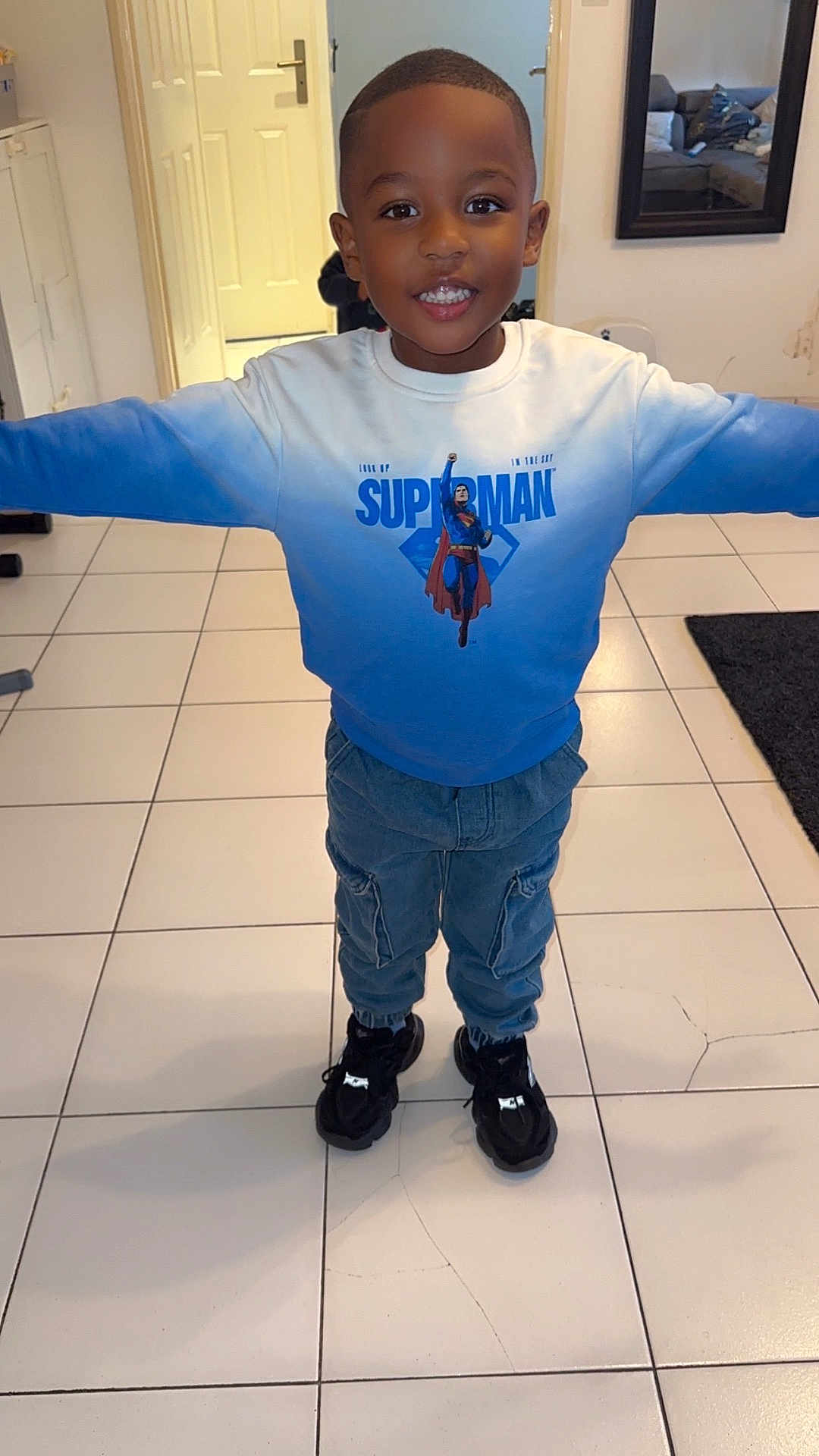 Imran a rejoint le concours — aidez-le/la à gagner de superbes lots ! child, boy, smiling, superman, sweatshirt, blue, pants, cargo_pants, black_shoes, indoor, tile_floor, door, mirror, room, happy, portrait, standing, casual_clothing, young, person