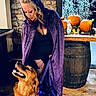 Daisy Mae is registered to the contest to win money with this photo: woman, dog, golden_retriever, purple_cloak, indoor, stone_wall, wooden_floor, pumpkins, barrel, decorations, leash, costume, harness, lights, smiling_dog, curly_hair, black_clothing, autumn, festive, looking_down