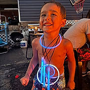 Braylen Yates is registered to the contest to win money with this photo: child, boy, smiling, glow_sticks, necklaces, shorts, shirtless, outdoor, evening, basketball_hoop, patio, furniture, happy, playful, summer, fun, light, joy, person, backyard
