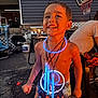 child, boy, smiling, glow_sticks, necklaces, shorts, shirtless, outdoor, evening, basketball_hoop, patio, furniture, happy, playful, summer, fun, light, joy, person, backyard