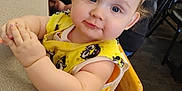 Phoenix is registered to the contest to win money with this photo: baby, child, high_chair, yellow_bib, table, indoor, person, face, blue_eyes, wooden_chair, clothing, cute, hands, seated, carpet, adult, background, skin, expression, hair