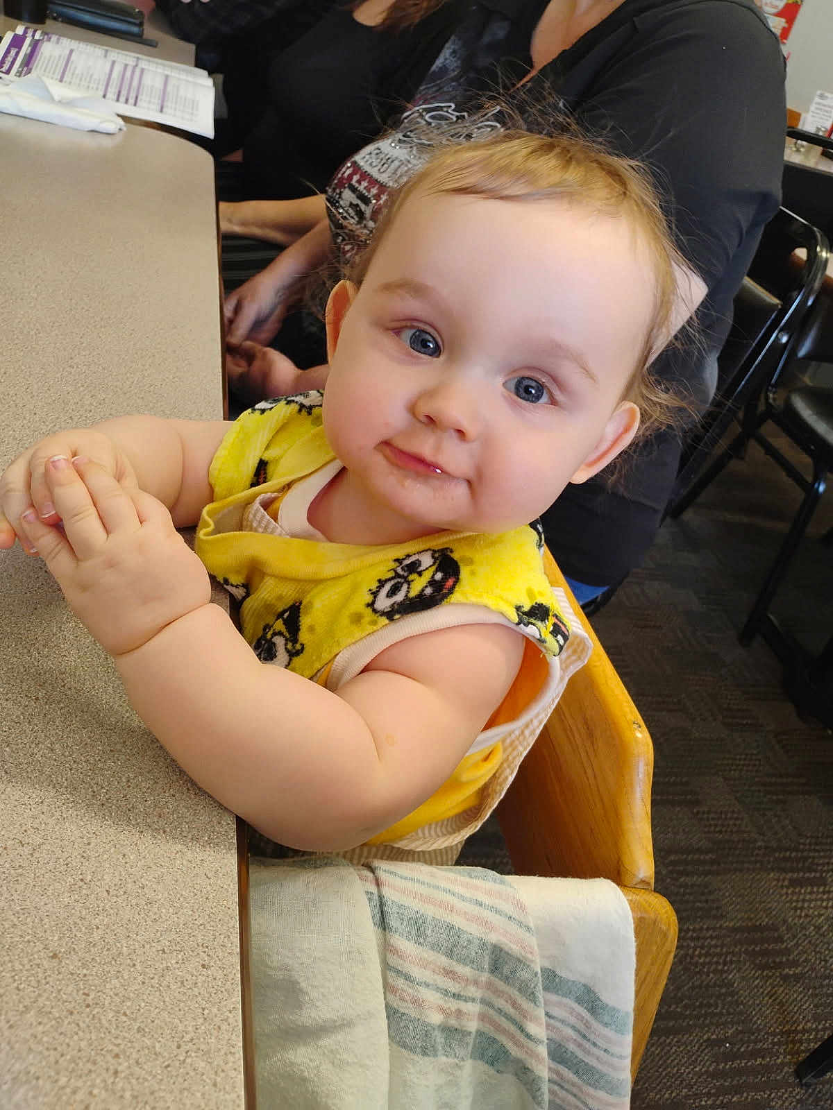 Phoenix is registered to the contest to win money with this photo: baby, child, high_chair, yellow_bib, table, indoor, person, face, blue_eyes, wooden_chair, clothing, cute, hands, seated, carpet, adult, background, skin, expression, hair