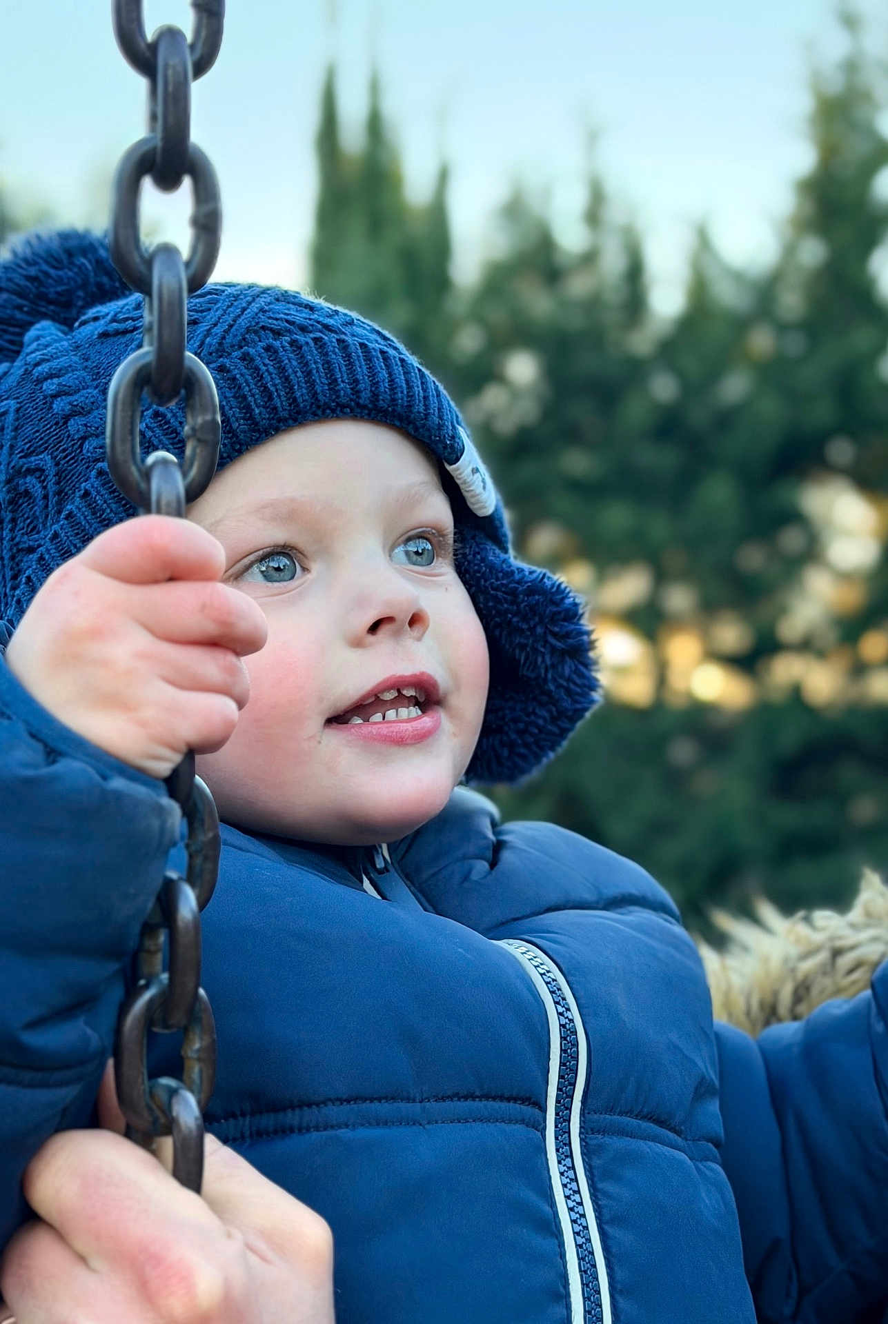 Aloïs participe au concours pour gagner de l'argent avec cette photo : child, blue_jacket, knitted_hat, swing_chain, outdoor, winter_clothing, close_up, face, hand, smiling, playground, nature, trees, cold_weather, happy, portrait, young, daylight, cute, expression