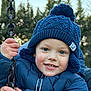 child, smiling, winter_clothing, blue_jacket, knitted_hat, pom_pom, swing, chain, outdoor, cold_weather, happy, portrait, face, person, nature, trees, daylight, cute, young_child, closeup