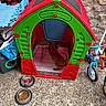 dog, doghouse, plastic, red, green, toy, tricycle, scooter, food_bowl, water_bowl, concrete, stone_wall, outdoor, pet, resting, blanket, playful, storage, colorful, relaxed