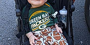 Ada is registered to the contest to win money with this photo: baby, child, stroller, green_bay_packers, football_pattern, bottle, clothing, smiling, outdoor, footwear, person, infant, happy, sitting, toy, black_stroller, casual, cute, young_child, baby_clothes