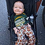 baby, child, stroller, green_bay_packers, football_pattern, bottle, clothing, smiling, outdoor, footwear, person, infant, happy, sitting, toy, black_stroller, casual, cute, young_child, baby_clothes
