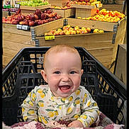 Ada is registered to the contest to win money with this photo: baby, shopping_cart, grocery_store, fruit, apples, produce_section, smile, child, infant, clothing, pattern, lemon, happy, retail, market, wooden_display, fresh, food, indoor, person