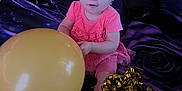 Amira is registered to the contest to win money with this photo: baby, child, pink_dress, headband, balloon, gold_ribbon, sitting, curious, indoors, blanket, foot, celebration, portrait, toddler, smile, face, person, cute, playing, happy