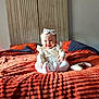 Elodie is registered to the contest to win money with this photo: baby, child, smiling, bed, bedspread, orange, headband, controller, toy, sitting, clothing, indoor, person, happy, cute, portrait, soft, warm, fabric, home