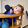 baby_bottle, blue_bib, blue_eyes, casual_clothing, child, curly_hair, cute, expression, face, hand, high_chair, indoor, infant, jeans, messy, playful, portrait, sitting, toddler, wooden_furniture