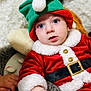 baby, child, festive_clothing, santa_outfit, elf_hat, blue_eyes, plush_toy, hand, red_clothing, white_fur_trim, gold_buttons, black_belt, soft_texture, indoor, portrait, cute, holiday, costume, infant, cozy