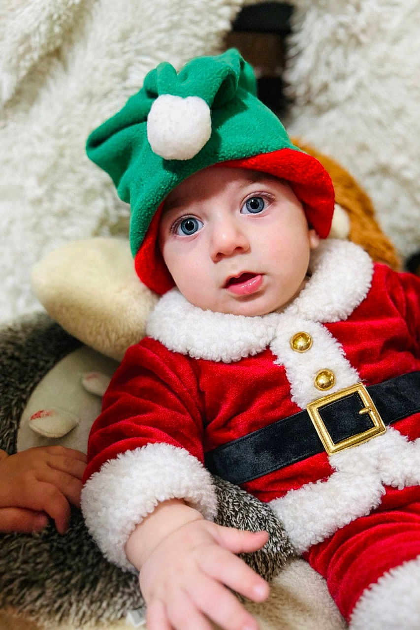 Omar participe au concours pour gagner de l'argent avec cette photo : baby, child, festive_clothing, santa_outfit, elf_hat, blue_eyes, plush_toy, hand, red_clothing, white_fur_trim, gold_buttons, black_belt, soft_texture, indoor, portrait, cute, holiday, costume, infant, cozy