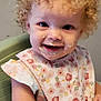 Kalia is registered to the contest to win money with this photo: toddler, child, curly_hair, high_chair, bib, food_on_face, smiling, cute, meal_time, indoor, baby, happy, portrait, eating, floral_pattern, small_child, messy, skin, person, face