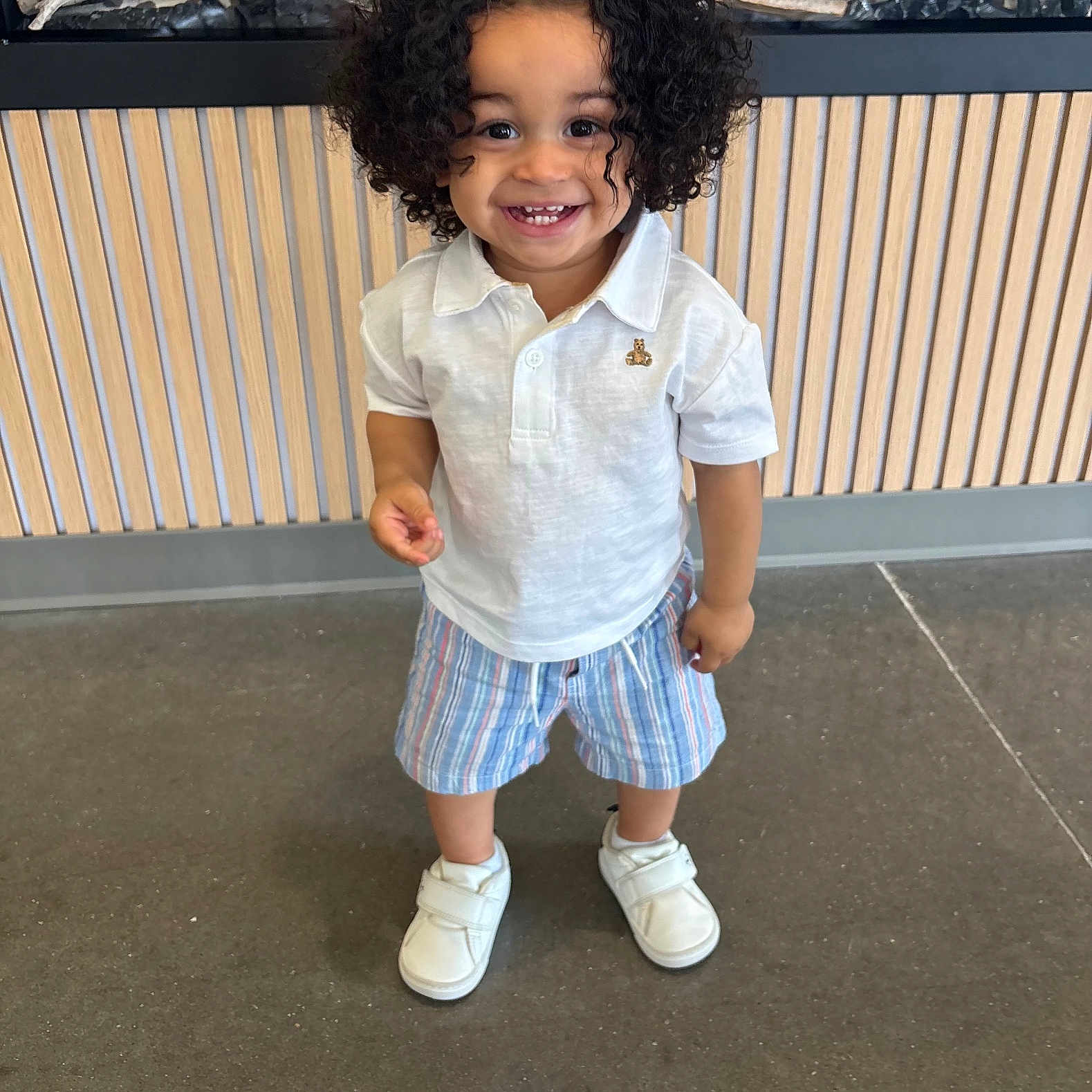 Namari joined the competition — help win amazing prizes! toddler, child, smiling, curly_hair, white_polo, striped_shorts, white_shoes, standing, indoor, floor, wooden_panel, decor, logs, happy, person, portrait, cute, clothing, young_child, feet