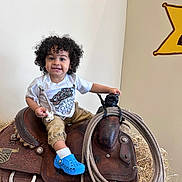 Namari is registered to the contest to win money with this photo: toddler, child, curly_hair, smile, saddle, lasso, rope, hay, blue_shoes, white_tshirt, khaki_pants, indoor, wall, sign, rustic, playful, happy, person, portrait, seated