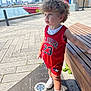 toddler, child, curly_hair, red_clothing, basketball_jersey, outdoor, bench, waterfront, city_skyline, boat, sunny, sidewalk, shoes, socks, urban, daytime, person, casual, standing, curious