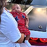 Maison is registered to the contest to win money with this photo: baby, child, person, holding, car, vehicle, red_clothing, striped_clothing, blue_stars, white_shirt, denim_shorts, outdoor, daylight, taillights, reflection, grass, trees, house, curious, casual