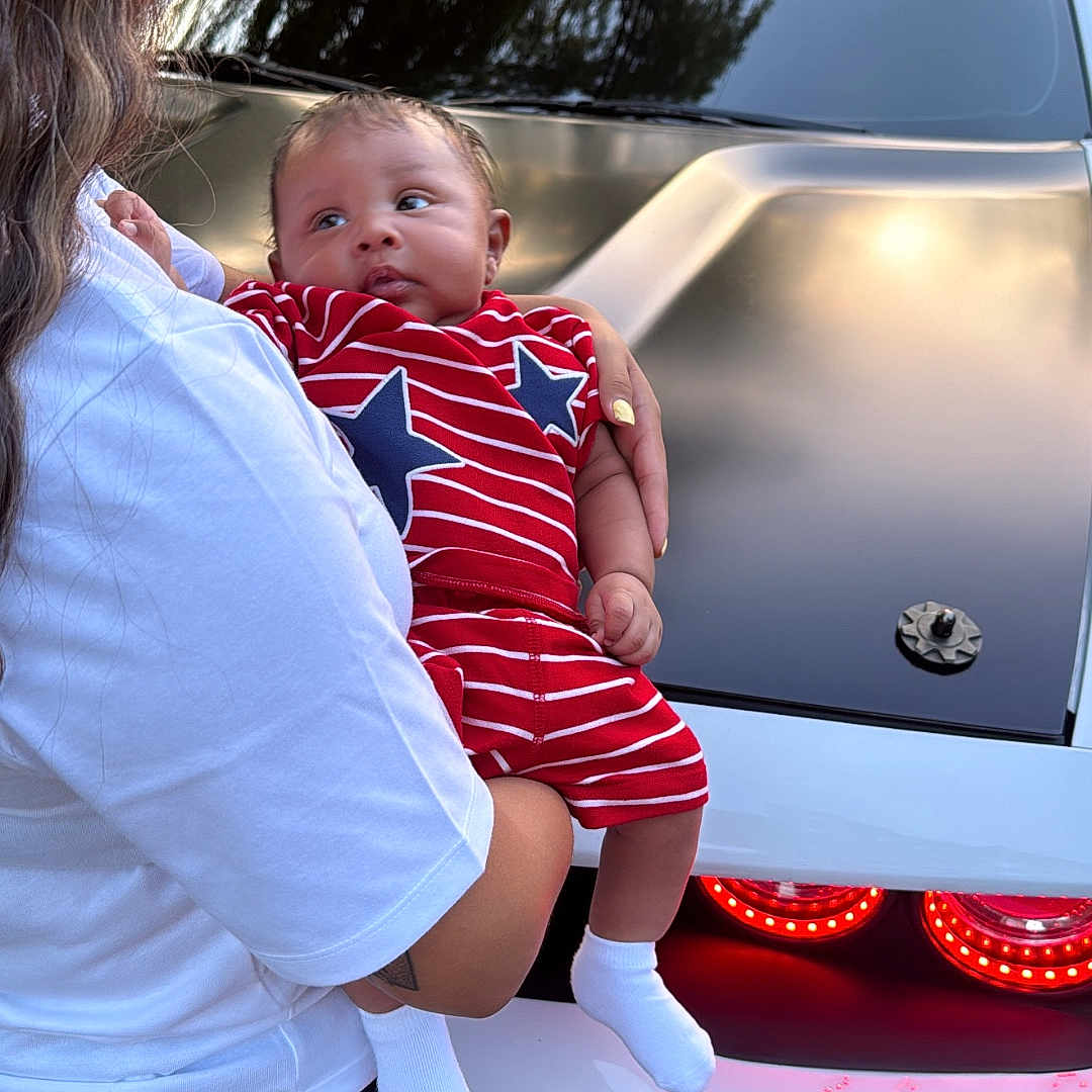 Maison is registered to the contest to win money with this photo: baby, child, person, holding, car, vehicle, red_clothing, striped_clothing, blue_stars, white_shirt, denim_shorts, outdoor, daylight, taillights, reflection, grass, trees, house, curious, casual