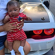 Maison joined the competition — help win amazing prizes! baby, child, person, holding, red_striped_outfit, star_pattern, yellow_nails, car, white_car, black_hood, tail_lights, road, trees, grass, outdoor, daylight, socks, legs, hand, casual_clothing