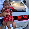baby, child, person, holding, red_striped_outfit, star_pattern, yellow_nails, car, white_car, black_hood, tail_lights, road, trees, grass, outdoor, daylight, socks, legs, hand, casual_clothing