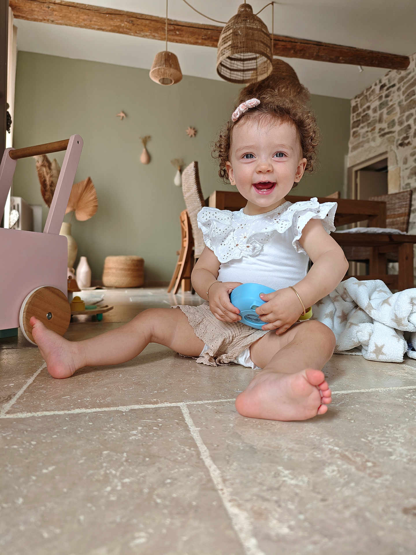 Margot participe au concours pour gagner de l'argent avec cette photo : toddler, child, smiling, curly_hair, toy, floor, indoor, wooden_furniture, wicker_lights, rustic_decor, cozy, playful, baby_clothes, hair_clip, natural_light, home, cute, happy, sitting, tiles