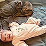 baby, cat, blanket, pink_clothing, headband, tortoiseshell_cat, indoor, smiling, lying_down, cozy, pet, infant, soft_texture, cute, resting, comfort, animal, human, face, portrait