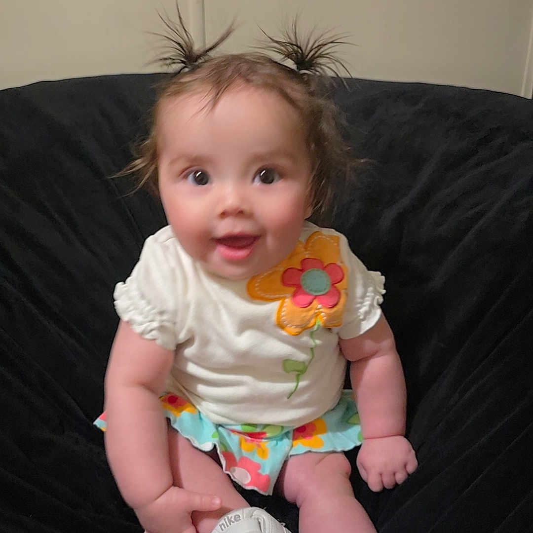 Birdie joined the competition — help win amazing prizes! baby, child, infant, sitting, smiling, white_shoes, flower_pattern, colorful_clothing, pigtails, cute, happy, indoors, black_chair, plush, person, young_child, casual, portrait, fun, playful