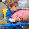 child, toddler, boy, sitting, shopping_cart, balloon, pink_balloon, parking_lot, cars, outdoor, daylight, blue_clothing, brown_hair, profile, holding, hugging, retail, supermarket, playful, portrait