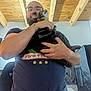 Freddy a rejoint le concours — aidez-le/la à gagner de superbes lots ! man, beard, glasses, cat, tabby_cat, holding, indoor, wood_ceiling, casual_clothing, tshirt, bracelet, jeans, affection, pet, home, furniture, chair, light, portrait, person