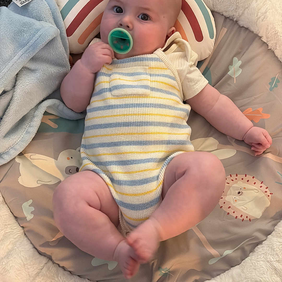 Jaxx-James is registered to the contest to win money with this photo: baby, blanket, carpet, child, chubby, clothing, comfort, cute, face, foot, hand, indoor, infant, lying_down, pacifier, plush_mat, rainbow_pillow, relaxed, soft_toys, striped_outfit