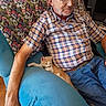 man, person, cat, kitten, orange_cat, white_cat, sitting, indoors, chair, armchair, plaid_shirt, glasses, bald, beard, blue_jeans, furniture, cozy, portrait, pet, relaxed