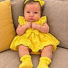 baby, infant, child, portrait, sitting, couch, yellow_dress, yellow_bow, headband, socks, smiling, facial_expression, indoors, furniture, cushion, bracelet, earrings, fashion, closeup, photography