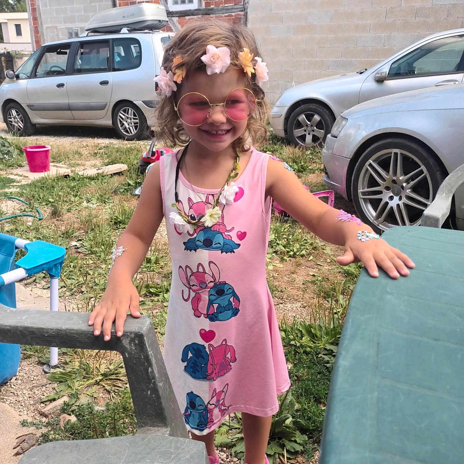Mya participe au concours pour gagner de l'argent avec cette photo : child, girl, pink_dress, sunglasses, flower_crown, outdoor, grass, cars, plastic_chair, plastic_table, smiling, cute, cartoon_print, accessories, summer, playful, happy, nature, casual, portrait