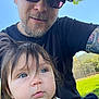 Olivia is registered to the contest to win money with this photo: child, toddler, man, adult, outdoor, park, grass, tree, sunlight, sunglasses, cap, beard, tattoo, face, portrait, nature, blue_sky, bench, fence, casual_clothing