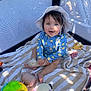 baby, child, playpen, hat, blue_clothing, toy, stuffed_animal, striped_towel, smiling, sitting, indoors, mesh_sides, sunlight, shadow, play_area, infant, cute, happy, colorful, young