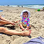baby, beach, sand, ocean, waves, sun_hat, colorful_clothing, child, sitting, legs, hands, sunny, daytime, outdoor, water, people, shore, relaxation, vacation, summer