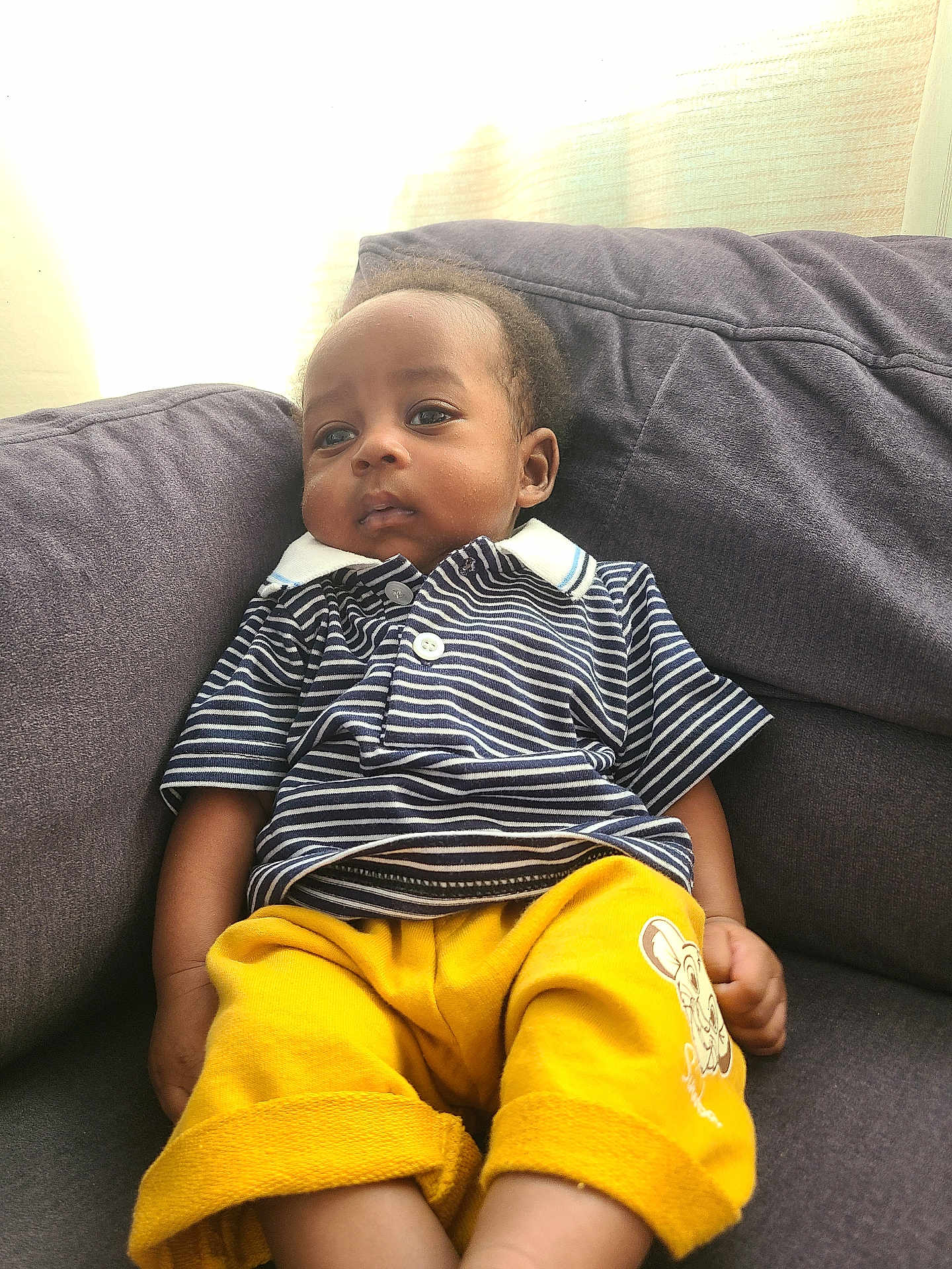 Kenny a rejoint le concours — aidez-le/la à gagner de superbes lots ! child, toddler, striped_shirt, yellow_shorts, couch, indoors, portrait, relaxed, casual_clothing, cartoon_print, natural_light, window_light, curtains, comfortable, young_child, seated, expression, home, soft_light, calm