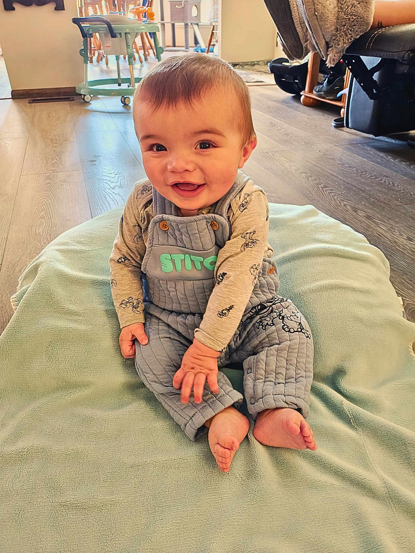 Silas is registered to the contest to win money with this photo: baby, child, smiling, sitting, blanket, overalls, long_sleeve, floor, wooden_floor, indoor, happy, person, face, feet, furniture, high_chair, home, cozy, cute, playful