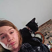 Sid joined the competition — help win amazing prizes! woman, cat, black_cat, pet, indoor, portrait, shoulder, collar, tag, clock, brick_wall, casual, expression, young_adult, plaid_shirt, face, feline, person, home, cute